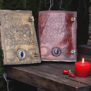 May include: Two leather-bound books titled "Book of Shadows" with embossed designs, including a pentagram and crescent moons. One book is tan, the other is reddish-brown. A red candle and decorative charms are also present.