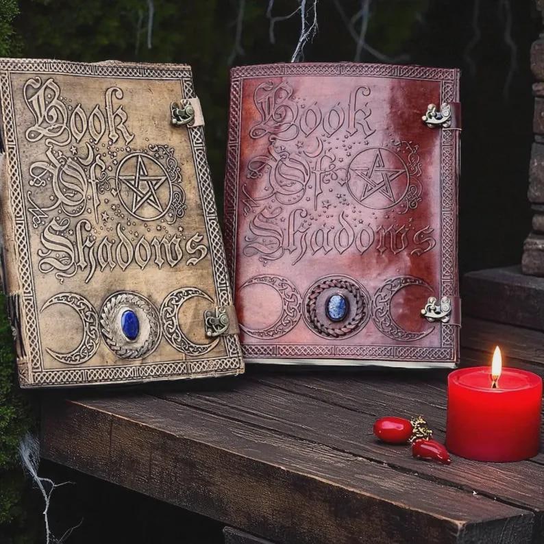 May include: Two leather-bound books titled "Book of Shadows" with embossed designs, including a pentagram and crescent moons. One book is tan, the other is reddish-brown. A red candle and decorative charms are also present.