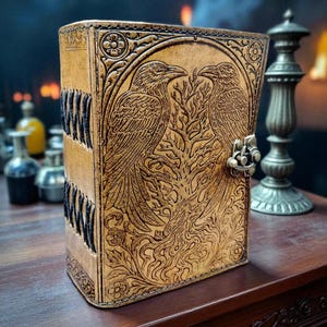 May include: A tan leather journal with a detailed embossed design featuring two ravens facing each other, a tree, and floral accents. The spine has black stitching. A metal clasp secures the journal.