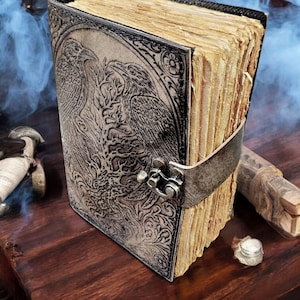 Puede incluir: Un diario encuadernado en cuero con un dise&ntilde;o de cuervo en relieve. El diario tiene un cierre de metal y una correa para mantenerlo cerrado. Las p&aacute;ginas est&aacute;n envejecidas, d&aacute;ndole un aspecto vintage. La cubierta es de color gris claro.