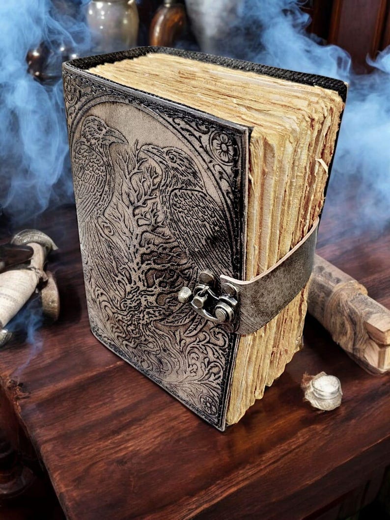 Puede incluir: Un diario encuadernado en cuero con un dise&ntilde;o de cuervo en relieve. El diario tiene un cierre de metal y una correa para mantenerlo cerrado. Las p&aacute;ginas est&aacute;n envejecidas, d&aacute;ndole un aspecto vintage. La cubierta es de color gris claro.