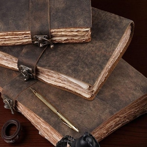 May include: Three brown leather journals with metal clasps and straps are stacked on a dark wooden surface. A gold pen and a small decorative item are next to the journals. The journals have aged, textured pages.