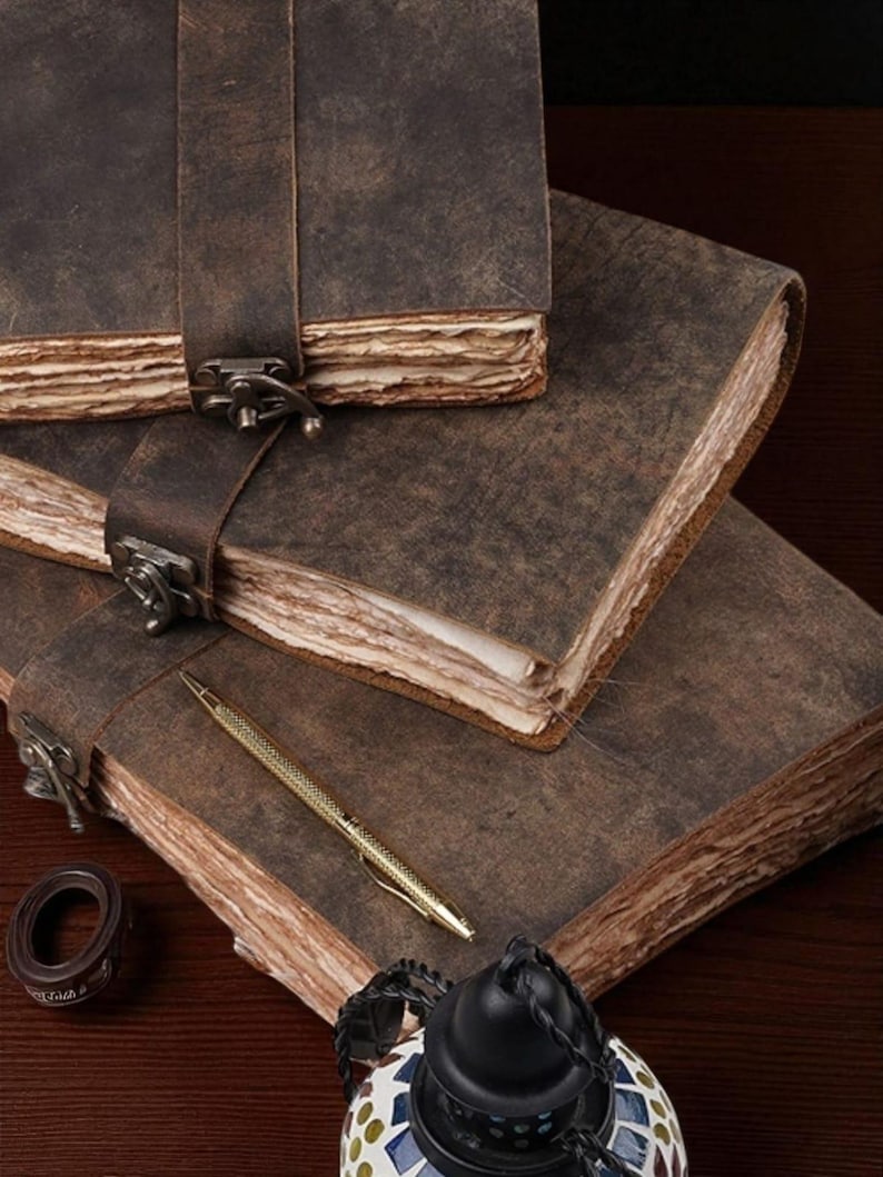 May include: Three brown leather journals with metal clasps and straps are stacked on a dark wooden surface. A gold pen and a small decorative item are next to the journals. The journals have aged, textured pages.
