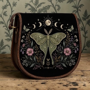 Könnte beinhalten: Eine schwarze Satteltasche mit braunem Lederbesatz, die auf der Vorderklappe eine zentrale Abbildung eines grünen Nachtfalters zeigt, umgeben von rosa Blumen, Blattwerk und einer Mondphasengrafik mit Vollmond und zunehmenden Mondsicheln.
