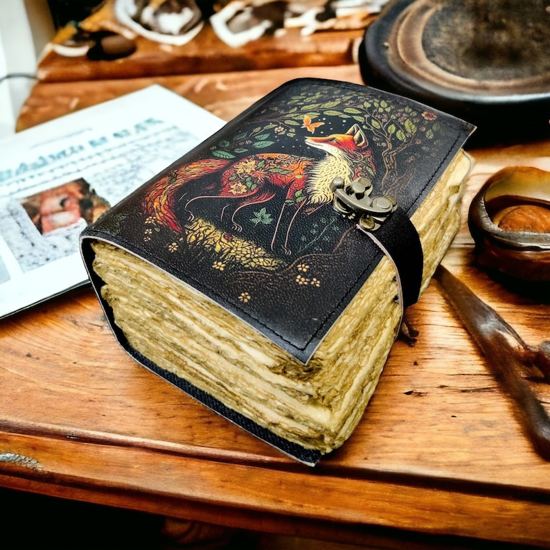 May include: A leather journal with a fox design on the cover. The fox is depicted in red, orange, and gold against a black background. The journal is secured with a black strap and metal clasp. The pages have a vintage, aged appearance, giving the journal a rustic feel.