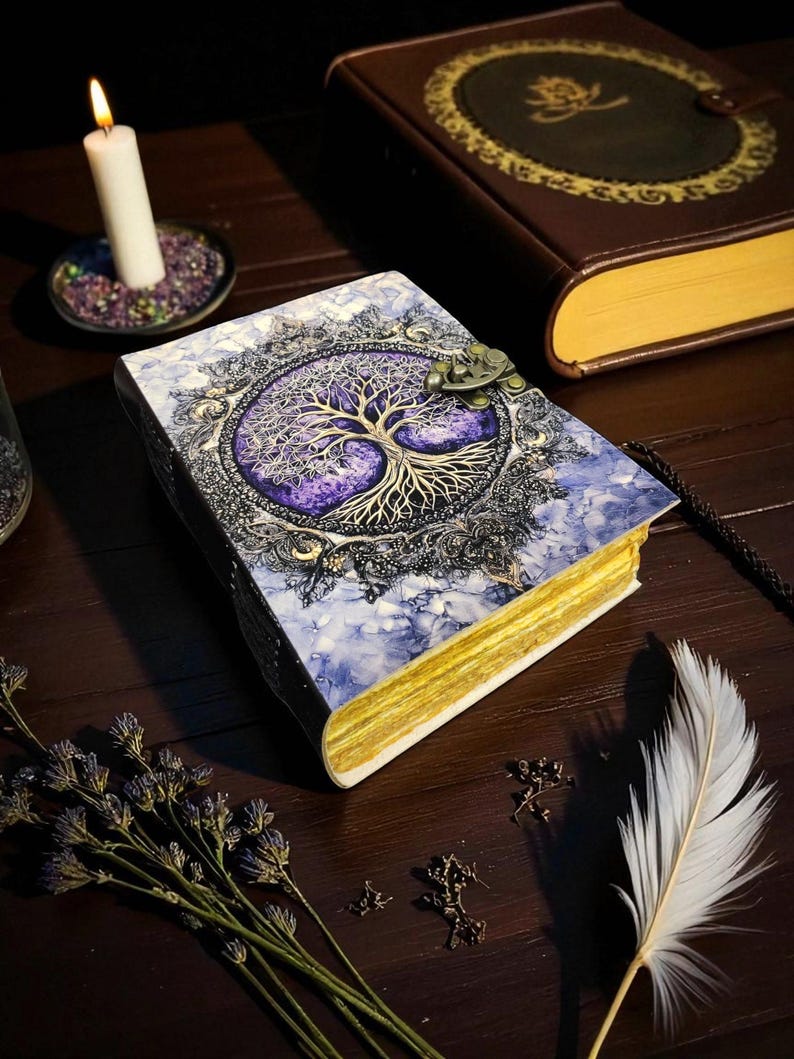 Op de afbeelding: Een decoratief boek met een levensboomontwerp op de kaft. De kaft is een mix van blauw en wit met een paars boomontwerp. Het boek heeft goudkleurige randen en een metalen sluiting. Een brandende kaars en een veer zijn ook zichtbaar.