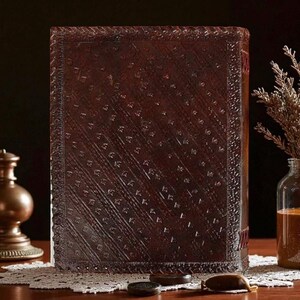 May include: A large, rectangular, dark brown leather-bound book with an embossed pattern. The book is standing upright on a wooden table, with a decorative lace doily and small objects in front of it. A glass jar and a brass lamp are visible in the background.