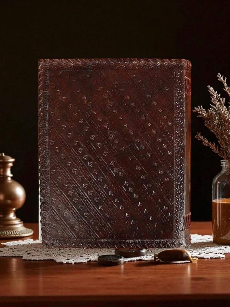 May include: A large, rectangular, dark brown leather-bound book with an embossed pattern. The book is standing upright on a wooden table, with a decorative lace doily and small objects in front of it. A glass jar and a brass lamp are visible in the background.