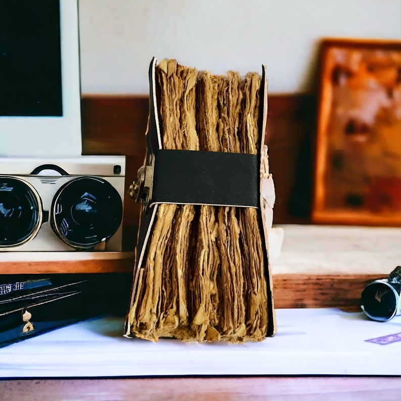 Puede incluir: Un diario de estilo vintage encuadernado en cuero con una correa de cuero marr&oacute;n oscuro y p&aacute;ginas de papel envejecido. El diario est&aacute; cerrado y se encuentra sobre una superficie blanca.