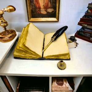 May include: An open book with aged, yellowed pages and a black cover. A black quill pen with gold detailing lies on the right page. An inkwell, a small gold candle holder, and a stack of books are also visible.