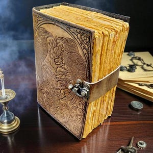 May include: A leather-bound journal with a detailed embossed design featuring two birds and floral patterns. The journal has aged, yellowed pages and a leather strap closure. The cover is a warm brown color, and the pages have a textured appearance.