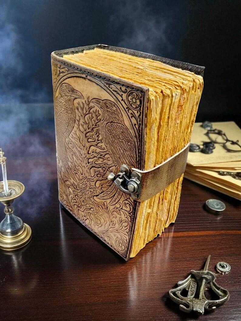 May include: A leather-bound journal with a detailed embossed design featuring two birds and floral patterns. The journal has aged, yellowed pages and a leather strap closure. The cover is a warm brown color, and the pages have a textured appearance.