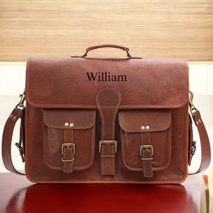 May include: A brown leather satchel with the name "William" embossed on the front. The bag features two front pockets with buckle closures, a top handle, and a shoulder strap. The bag is rectangular and has a vintage look.