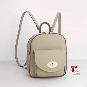 May include: A beige leather backpack with a rounded shape, featuring a front pocket and a gold-colored clasp. The bag has a top handle and adjustable shoulder straps. White stitching outlines the edges of the bag and pocket.