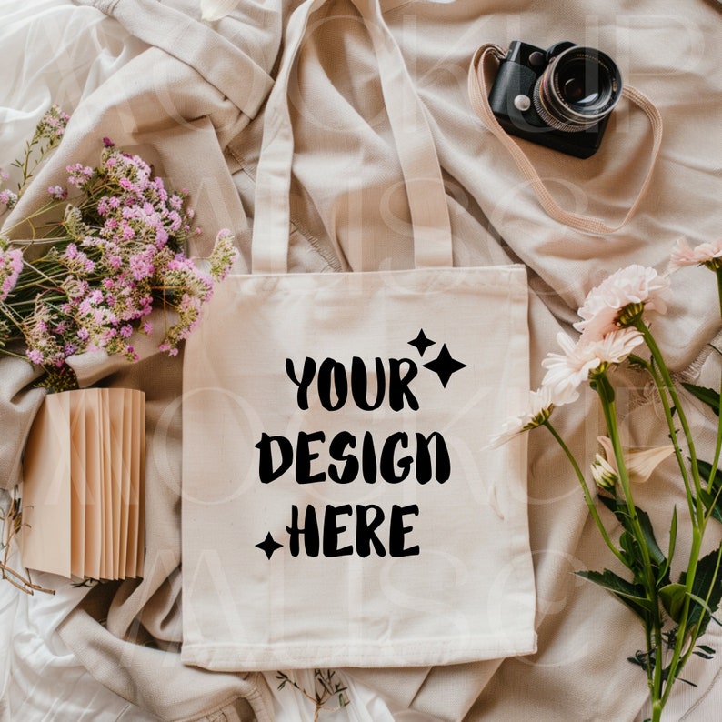 May include: A white canvas tote bag with black handles and the text "YOUR DESIGN HERE" printed on the front. The bag is lying on a beige fabric surface with flowers and a camera.