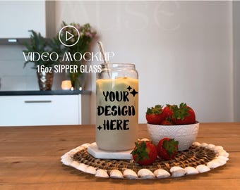 May include: A 16oz sipper glass mockup with a straw, filled with a light brown liquid. The glass has the text "Your Design Here" printed on it. The glass is sitting on a woven placemat with a bowl of strawberries in the background.