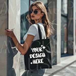 May include: A woman wearing sunglasses and a white t-shirt is holding a black tote bag with the text "YOUR DESIGN HERE" printed on it.