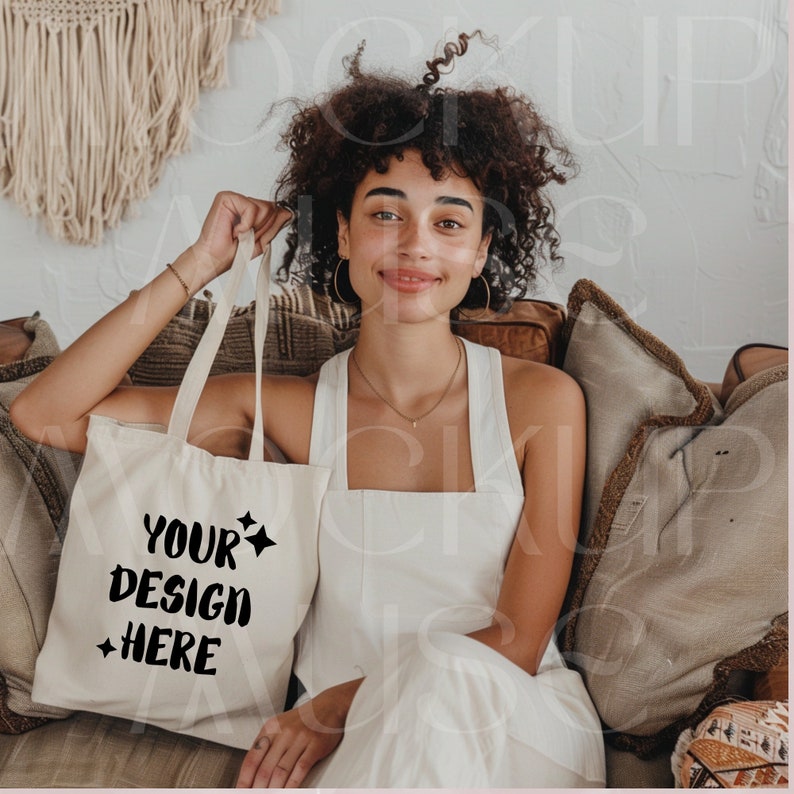 Może przedstawiać: Kobieta siedząca na kanapie trzymająca białą torbę typu tote z napisem "YOUR DESIGN HERE" w kolorze czarnym. Torba ma po obu stronach napisu czarny wz&oacute;r gwiazdy.