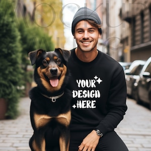 May include: A man wearing a black sweatshirt with the text "YOUR DESIGN HERE" in white, a black beanie, and black pants is smiling and looking at the camera. He is standing next to a black and brown dog.