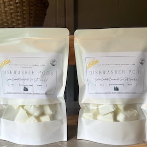 Op de afbeelding: Twee witte zakken met vaatwastabletten met een label waarop "Sweet Lemon Dishwasher Pods" en "Non-Toxic Safe Gentle Squeaky Clean" staat. De tabletten zijn gemaakt van soda, boorax en zeezout-citroenolie. De zakken liggen op een houten oppervlak.