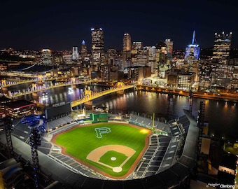 May include: An aerial night view of a baseball stadium with a city skyline in the background. The stadium is lit up and the field is green. The city skyline is also lit up and includes tall buildings.