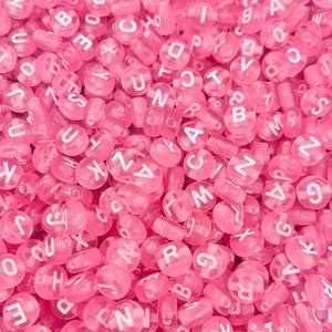 May include: A large assortment of pink alphabet beads. The beads are made of plastic and have white letters on them.