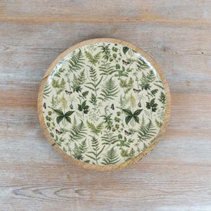 Könnte beinhalten: Ein rundes Holztablett mit einer cremefarbenen Oberfläche, die mit einem wiederkehrenden Muster aus verschiedenen grünen Farnblättern und kleinen braunen Schmetterlingen verziert ist. Das Tablett hat einen Rand aus hellem Naturholz.