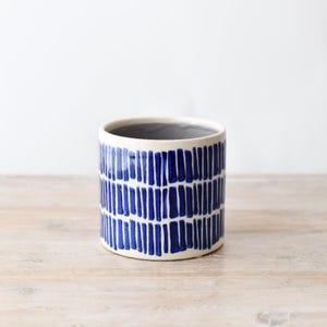 Op de afbeelding: Cilindrische keramische plantenbak met een crèmekleurige buitenkant en een blauw verticaal strepenpatroon. De binnenkant is grijs. De plantenbak staat op een lichtgekleurd houten oppervlak. Ideaal voor kleine planten of vetplanten.