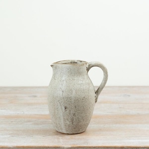 May include: A ceramic pitcher with a speckled off-white and light grey finish. The vessel features a rounded body, a curved handle on the right side, and a small pouring spout at the top rim. The surface has a textured, matte appearance.