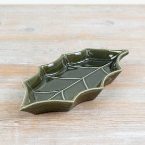 Può includere: Un piatto in ceramica verde oliva scuro lucido a forma di foglia appuntita. Il piatto presenta una superficie liscia e riflettente con linee incavate che imitano le venature di una foglia, creando una forma organica con bordi ricurvi.