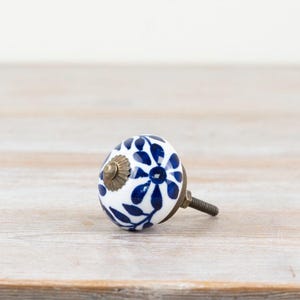 Könnte beinhalten: Ein weißer Keramik-Schrankknauf mit blauem Blumenmuster. Der Knauf hat einen messingfarbenen Sockel und eine Gewindeschraube zur Befestigung. Das Design zeigt eine zentrale Blume und Blätter.