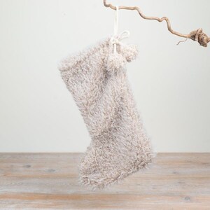 May include: A beige and white fuzzy Christmas stocking made of soft, shaggy yarn. The stocking features a long, textured pile and is accented with two small, matching pom-poms attached to a white cord loop at the top for hanging purposes.