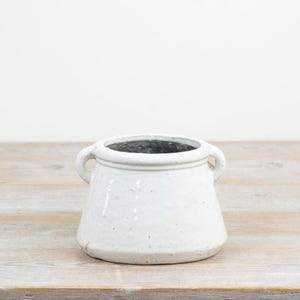 May include: A white ceramic pot with a wide, tapered base and a thick, rounded rim. The vessel features two small, integrated side handles and a smooth, glossy finish with subtle, natural speckling throughout the exterior surface.