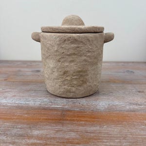 May include: A cylindrical beige stoneware jar with a rough, textured surface, featuring two small side handles and a matching circular lid with a central knob. The container has a neutral, earthy tone and a consistent, granular appearance throughout.