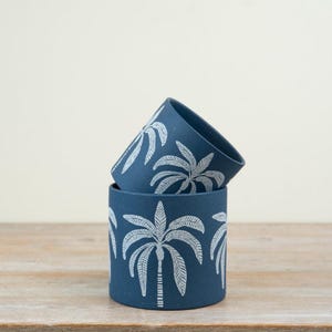 May include: Two navy blue cylindrical planters with white palm tree designs. One planter is stacked on top of the other. The planters are made of a matte material and sit on a wooden surface.