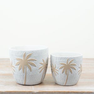 Op de afbeelding: Twee witte cilindrische plantenbakken met een getextureerd verticaal geribbeld oppervlak en een herhalend patroon van lichtbruine palmbomen in reliëf aan de buitenkant. De plantenbakken hebben verschillende formaten.