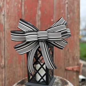 May include: A black metal lantern with a white candle inside, topped with a large black and white striped bow. The lantern has a geometric design and sits on a weathered metal surface. The background features a red wooden door.