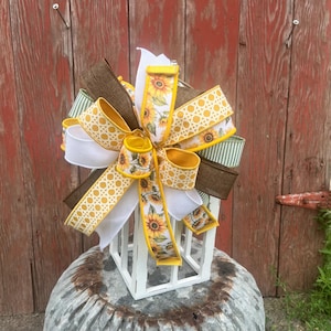 May include: A decorative bow with multiple ribbons in various patterns and colours. The bow includes ribbons with sunflower designs, white, brown, and yellow lattice patterns, and green and white stripes. The bow is placed on top of a white, square lantern.
