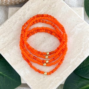 May include: A stack of four circular bracelets made of small, vibrant orange seed beads. Each bracelet features a single, small, polished gold-toned metallic bead integrated into the strand of orange beads, creating a uniform and consistent design.