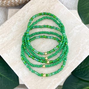 May include: A stack of five circular bracelets made of small green seed beads. Each bracelet features a single, small, polished gold-tone metal bead integrated into the strand of green beads, creating a uniform and consistent design.