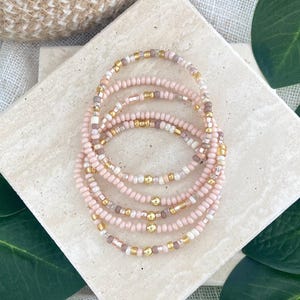May include: A stack of five beaded bracelets featuring small, round, light pink beads interspersed with metallic gold-toned spacer beads. The bracelets are arranged in a circular formation, showcasing a consistent pattern of pink and gold elements throughout each individual strand.