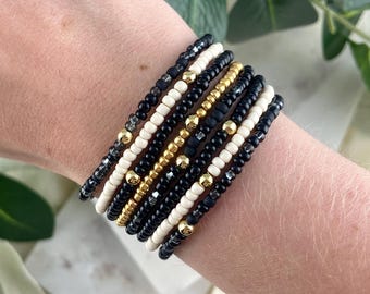 May include: A stack of seven beaded bracelets featuring a repeating pattern of small black, cream, and metallic gold beads. The bracelets are arranged in a uniform row, alternating between matte black, smooth cream, and shiny gold spherical accents.