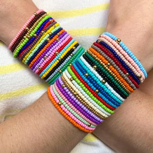 May include: A collection of stacked bracelets featuring small, colorful glass beads in various hues including pink, blue, green, yellow, and orange, interspersed with small, reflective gold-toned metallic spacer beads throughout the designs.