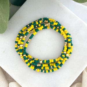 May include: A set of beaded bracelets in a spiral arrangement. The bracelets feature a pattern of yellow and green beads, interspersed with gold-colored beads. The bracelets are displayed on a white surface with a hint of greenery in the background.