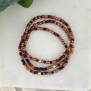 May include: Three beaded bracelets in shades of brown, copper, and gold. The bracelets are made of small, round beads and feature gold-colored accents. The bracelets are arranged in a circular pattern on a white surface.