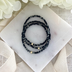 May include: Three beaded bracelets in black and gray with gold accents. The bracelets are made of small, round beads and are arranged in a circular pattern. The bracelets are displayed on a white marble surface.