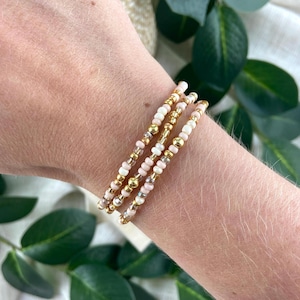 May include: A set of three delicate beaded bracelets featuring a combination of small round pale pink beads, metallic gold-tone beads, and clear translucent beads arranged in a repeating pattern around each elasticized strand.