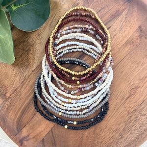 May include: A collection of beaded bracelets in various colors, including black, white, gold, and burgundy. The bracelets are arranged in a circular pattern on a wooden surface, showcasing their diverse designs and textures. The beads appear to be small and uniform in size.