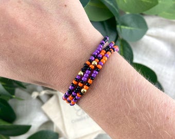 May include: A multi-strand bracelet featuring small, round beads in alternating colors of purple, orange, and black. The design includes several small, shiny gold-toned metallic spacer beads interspersed throughout the beaded strands.