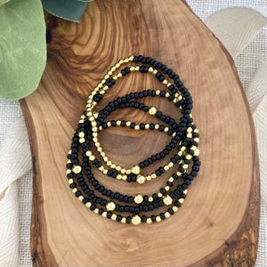 May include: A set of beaded bracelets featuring black and gold beads. The bracelets are arranged in a stack, showcasing the contrast between the dark and shiny elements. The bracelets are displayed on a wooden surface.